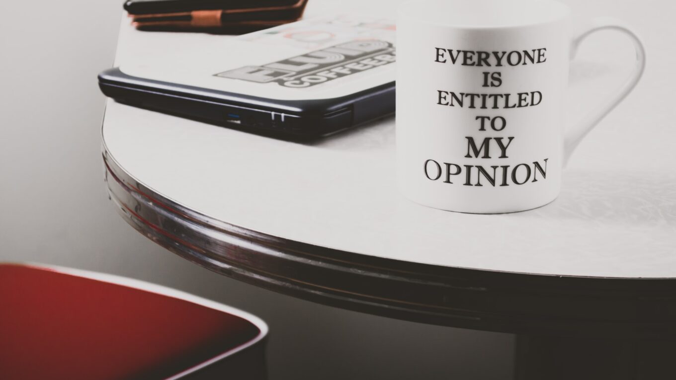 White mug on the edge of a table, with 'everyone is entitled to my opinion' wirtten on it