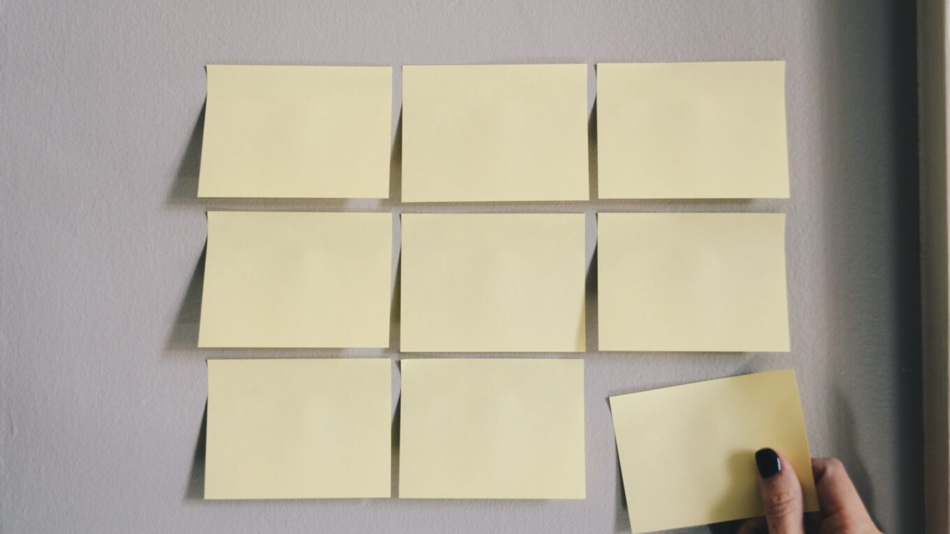 A 3x3 grid of 9 post-it notes on the wall, the lower right one being removed by a female hand